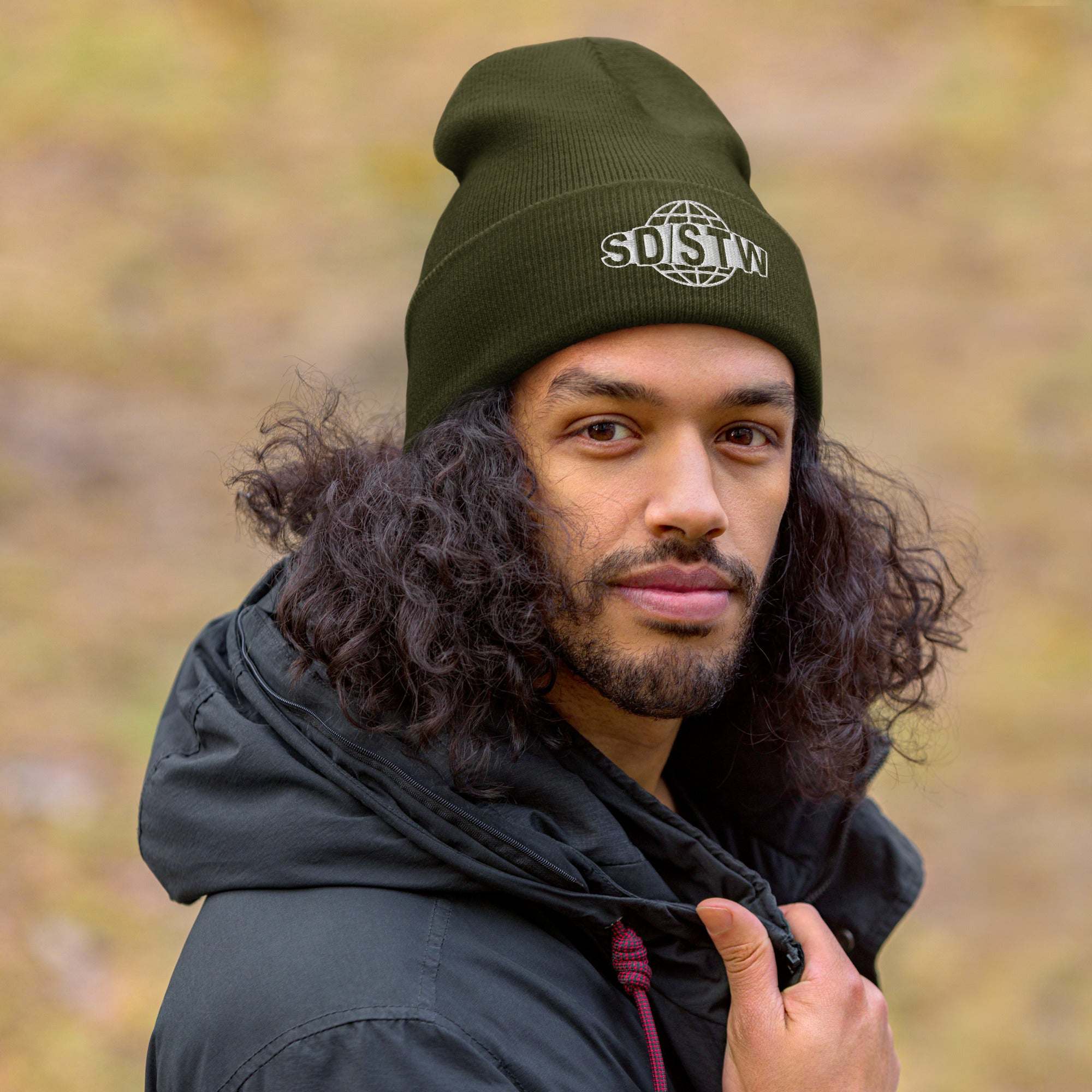 Man wearing olive green Suck Dick Save the World beanie with embroidered SD/STW logo outdoors