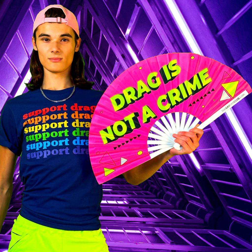 Person holding a UV-reactive drag hand fan with neon "Drag Is Not A Crime" text and colorful background