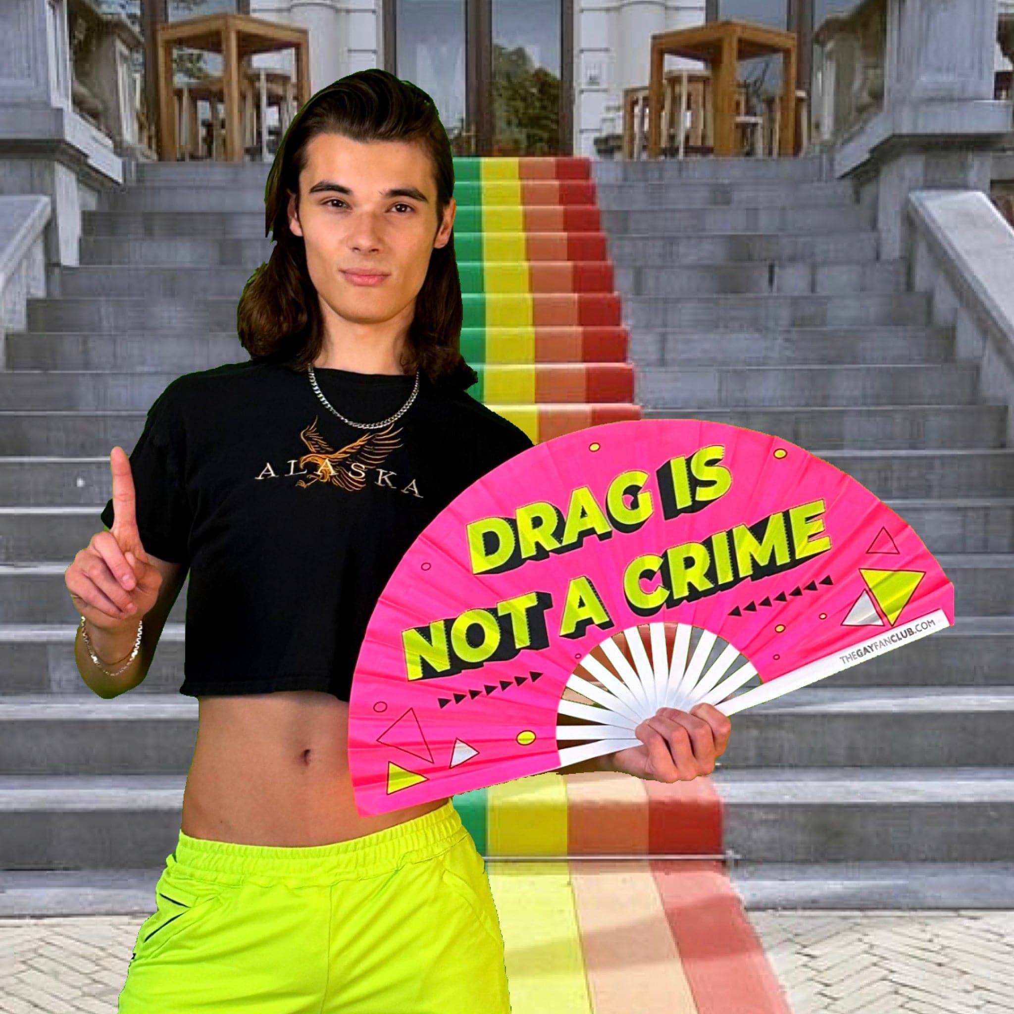 Person holding UV-reactive drag hand fan with neon colors and "Drag Is Not a Crime" text in front of rainbow stairs