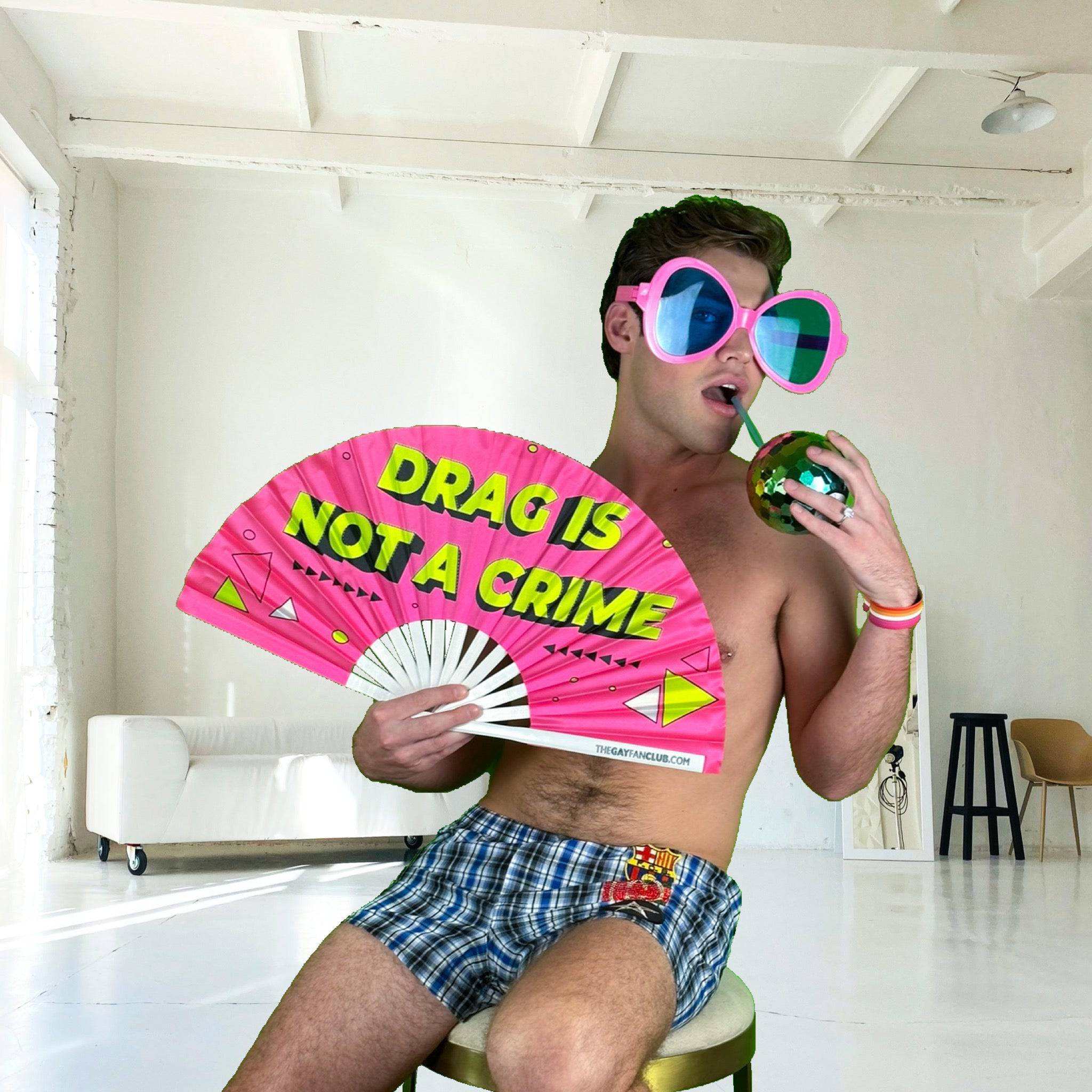 UV-reactive drag hand fan with neon "Drag Is Not A Crime" print held by person in oversized sunglasses