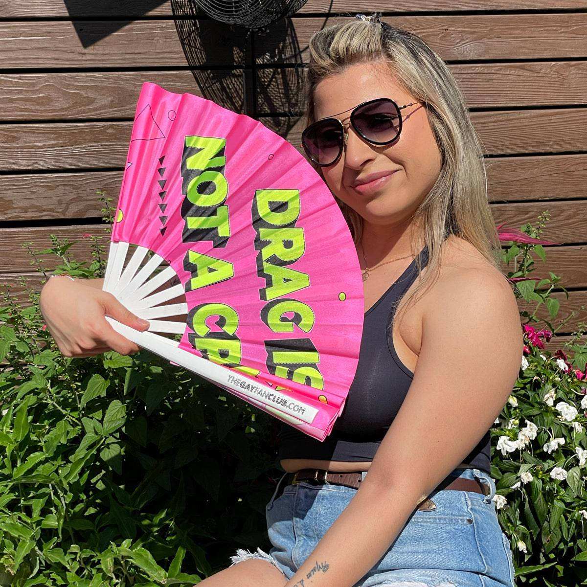 Woman holding pink UV-reactive drag hand fan with neon "Drag Is Not A Crime" text outdoors