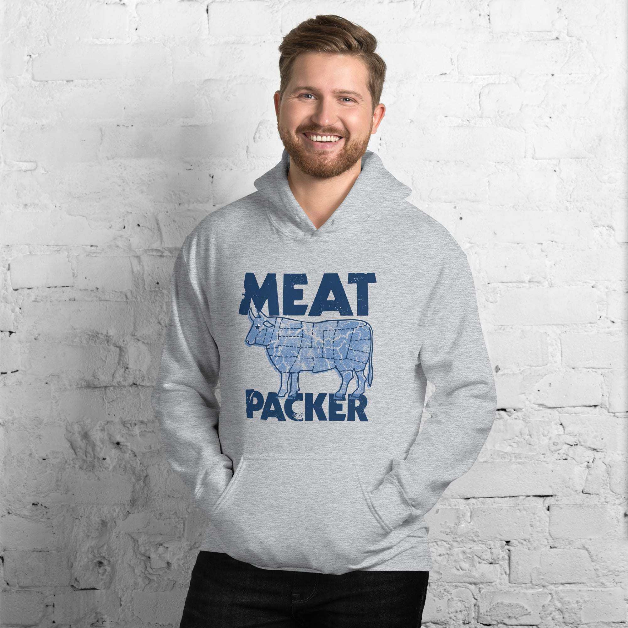 Man wearing grey Meat Packer Pullover Hoodie with blue graphic and text, standing against white brick wall